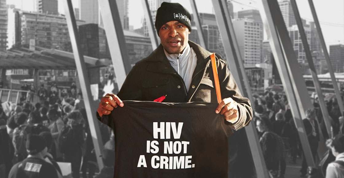 Man standing in front of a grayscale photograph holding a black t-shirt that says "HIV is not a crime" in white text